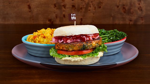 A soft roll with a chickpea and spinach patty, houmous, red pepper chutney and pink pickled onions on a plate with long-stem broccoli and spicy rice.